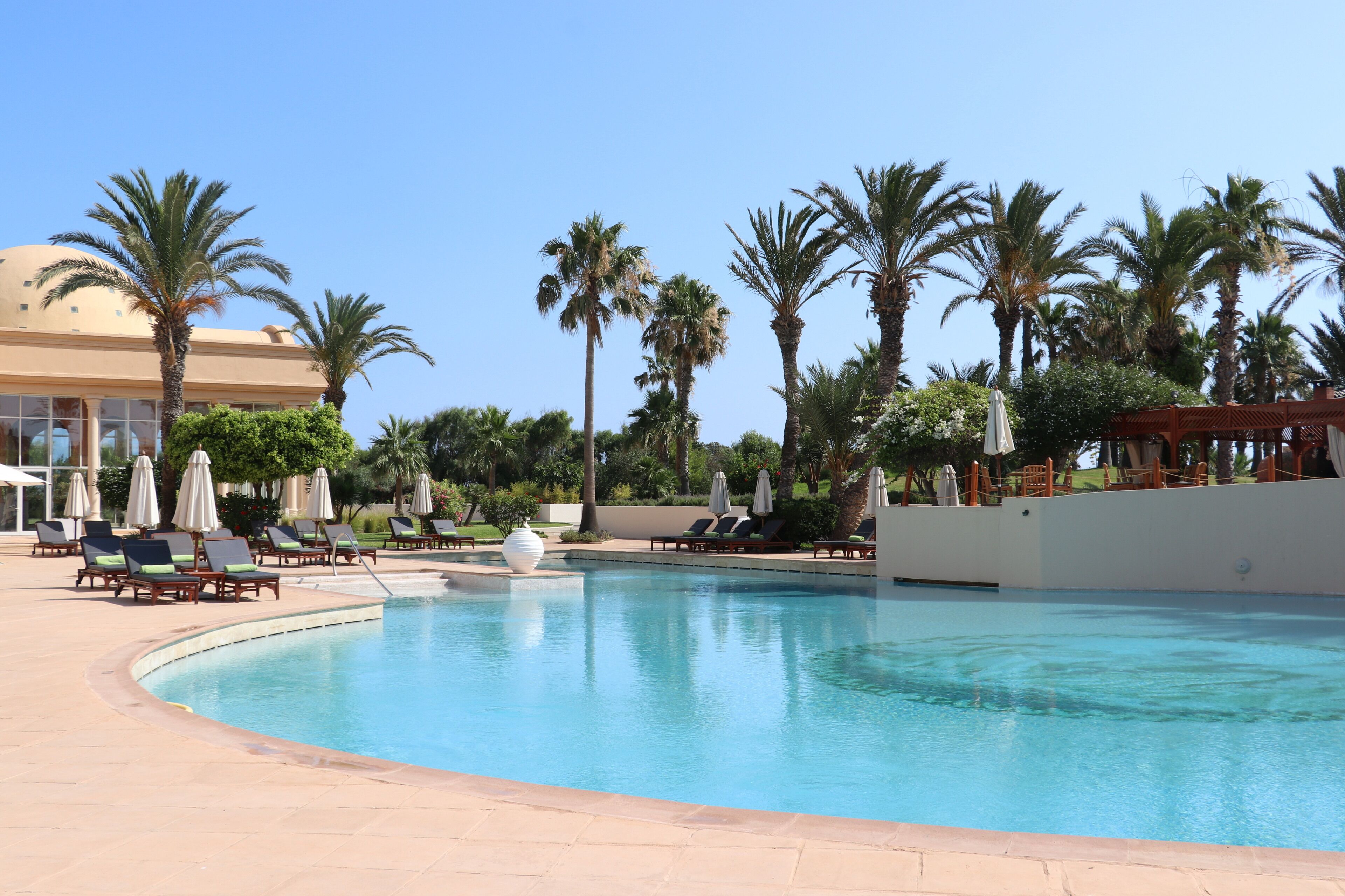 A pool area with lounge chairs and umbrellas, surrounded by palm trees and a building with a dome.