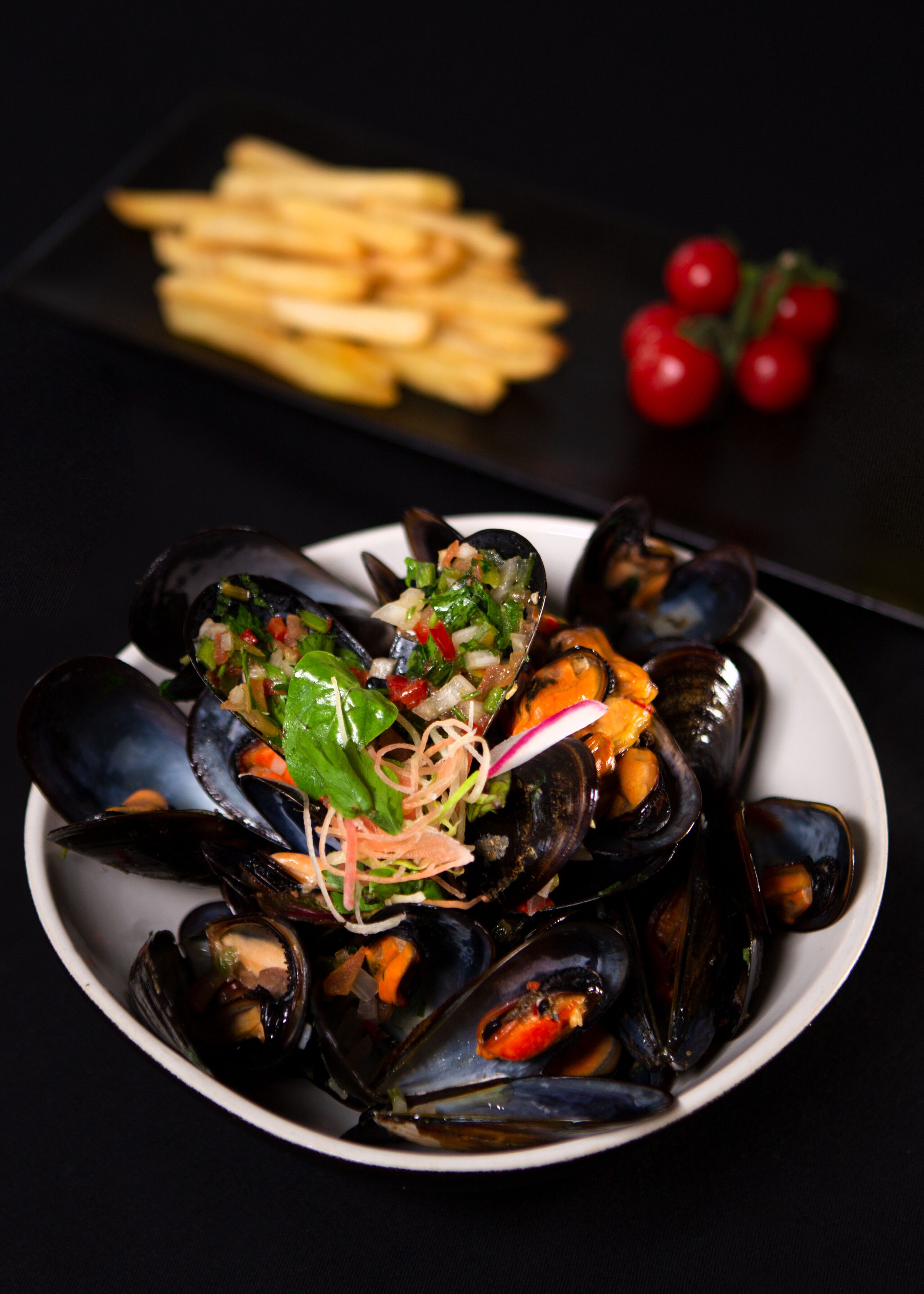A plate of mussels with a garnish of vegetables and herbs, accompanied by a side of fries and cherry tomatoes on a dark background.
