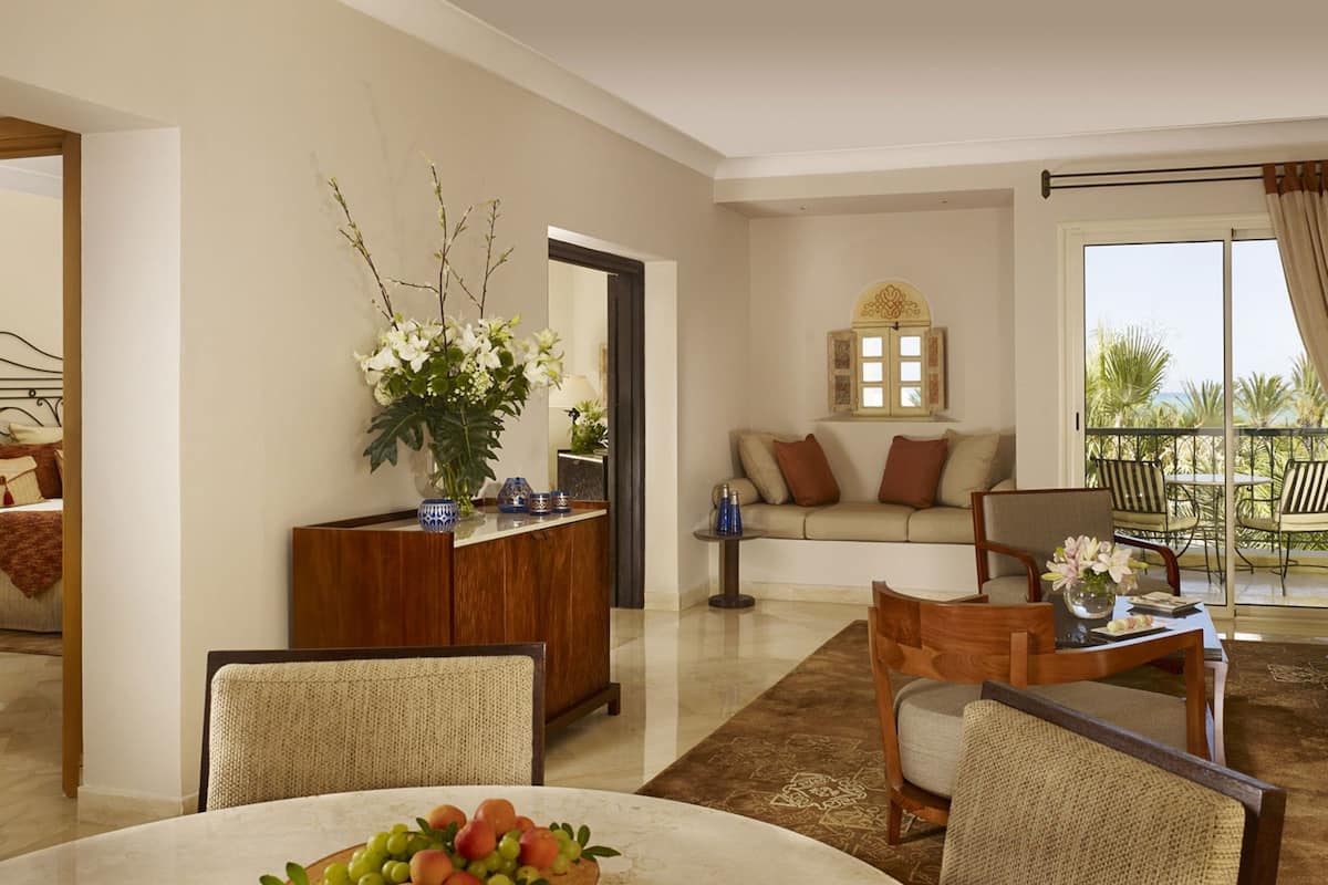 A living room with a sofa, chairs, a coffee table, and a dining table with fruit. There is a decorative vase with flowers and a mirror on the wall.