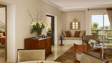 Una sala de estar con sofá, sillas, una mesita de centro y una mesa de comedor con fruta. Hay un jarrón decorativo con flores y un espejo en la pared.