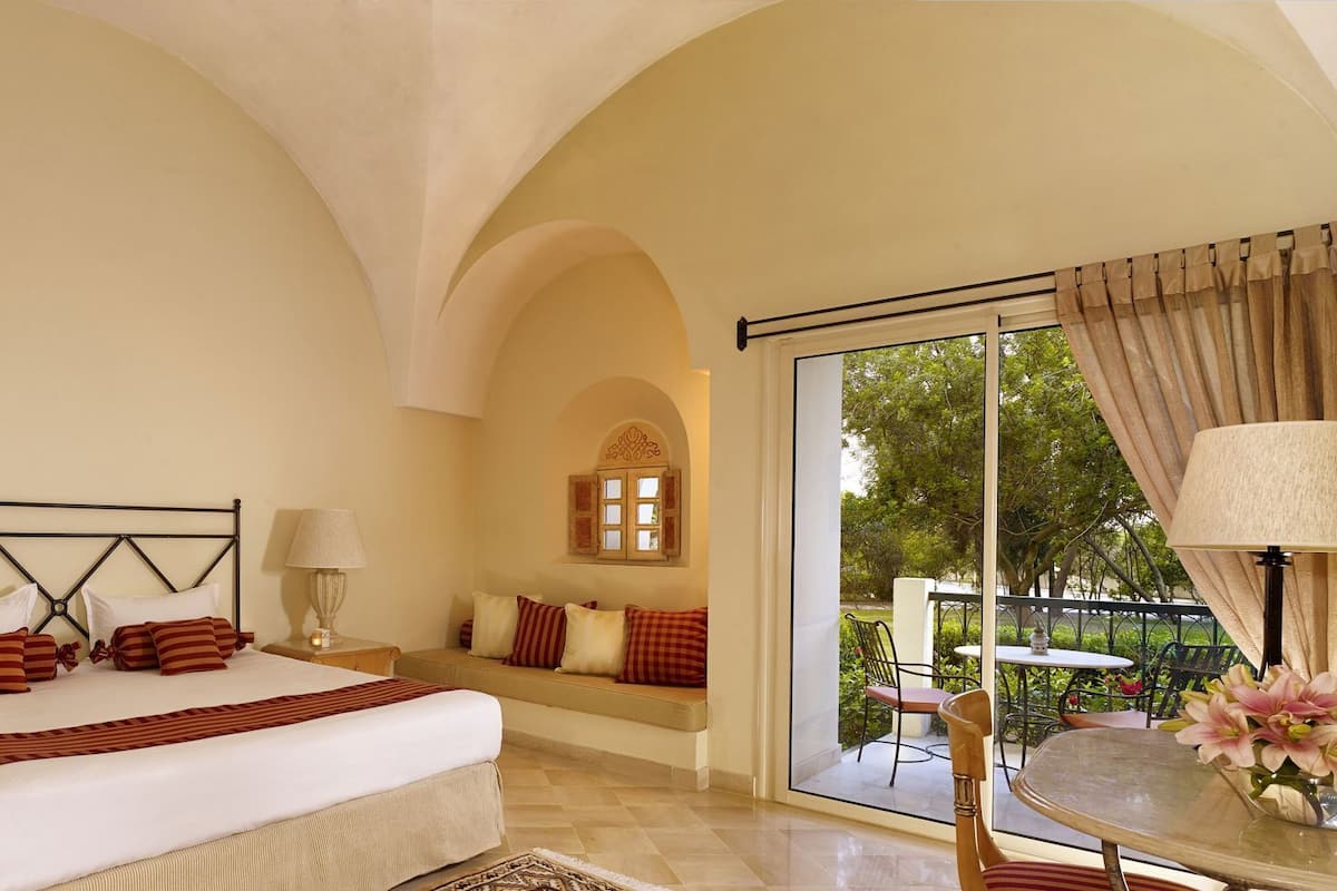 A bedroom with a bed, bedside lamps, a bench with cushions, a window, and a view of the outdoors.