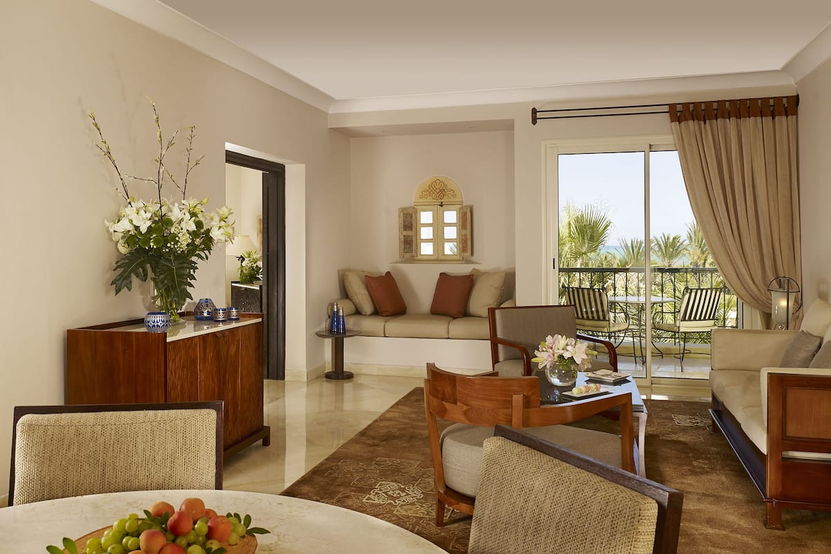 A spacious living room with a sofa, chairs, a dining table with fruit, a wooden cabinet, and a balcony with a view.