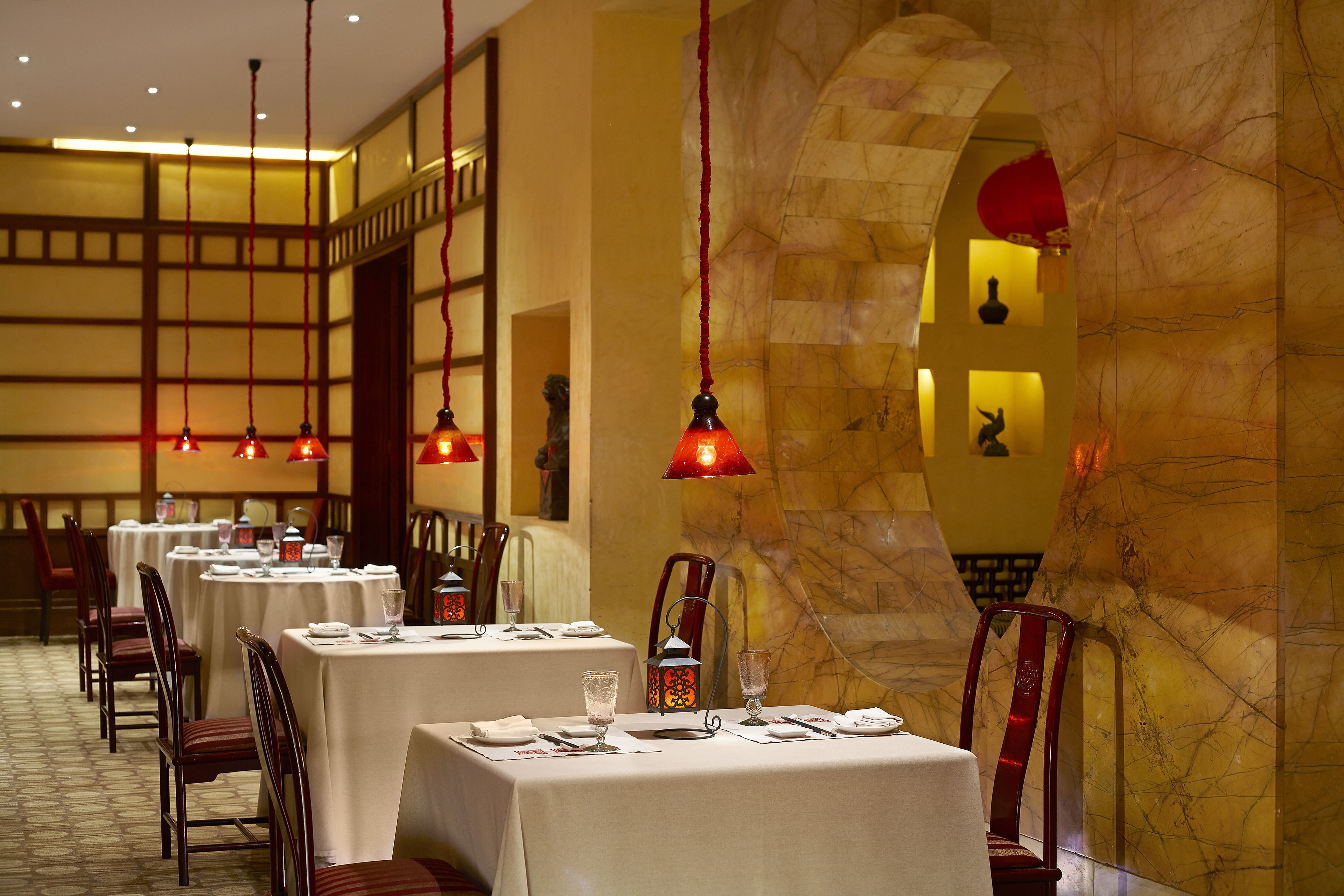 Un restaurante con varias mesas, lámparas rojas colgantes y una pared de mármol.