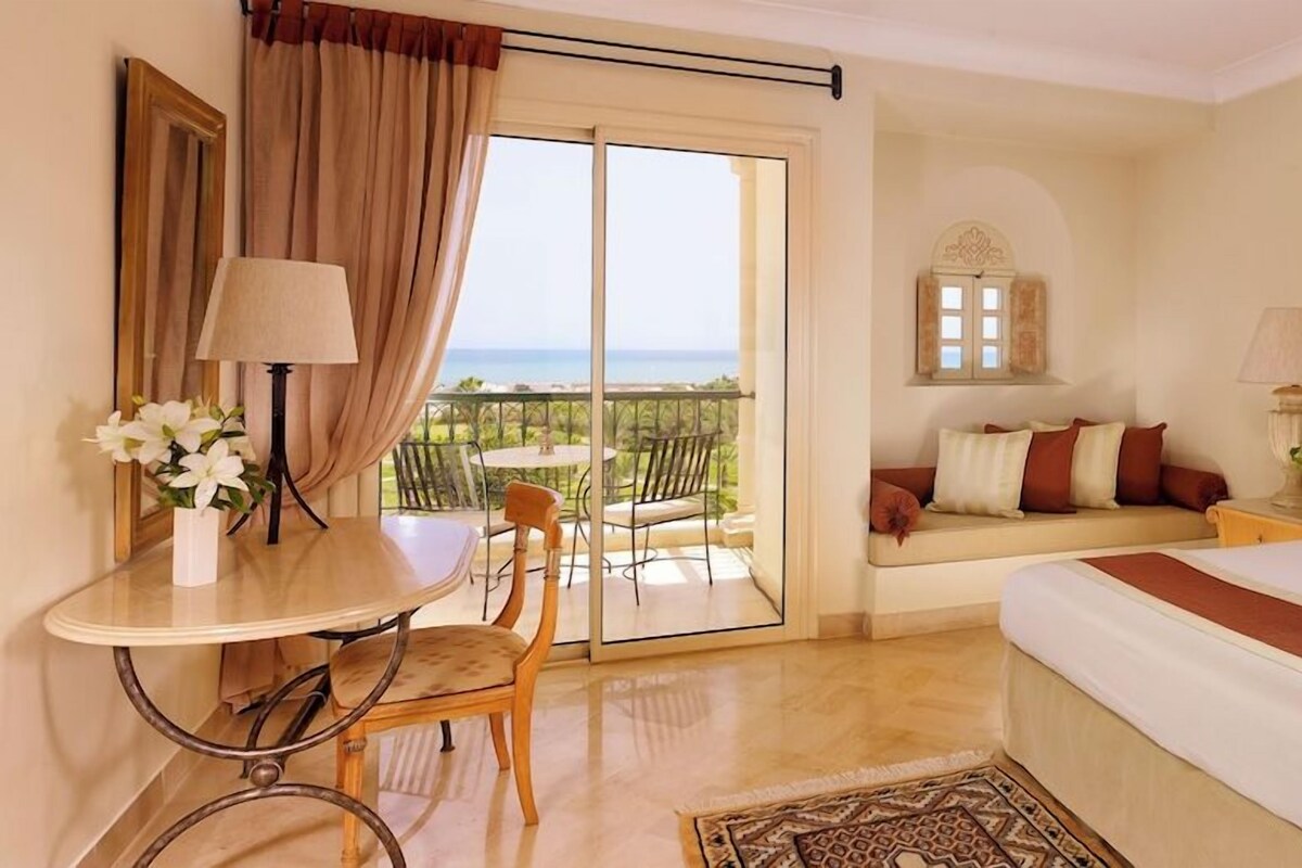 A hotel room with a bed, a sofa, a table with a lamp, a chair, and a view of the sea through a sliding door.