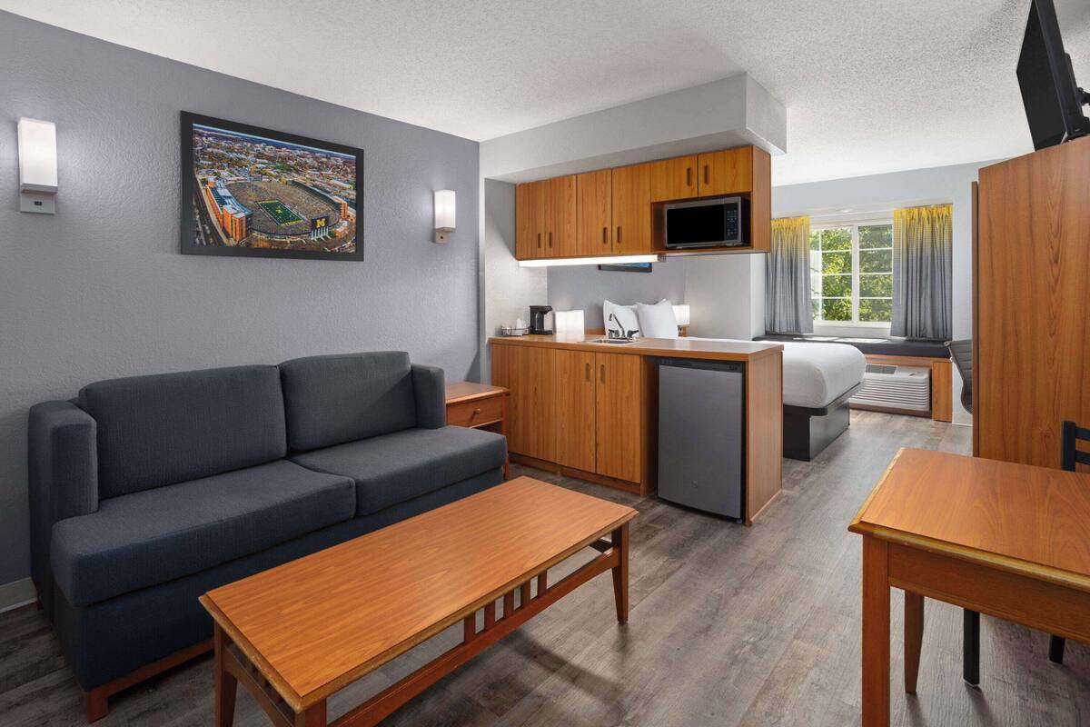 A compact hotel room with a sofa, a wooden table, a kitchenette, a bed, and a window with curtains.