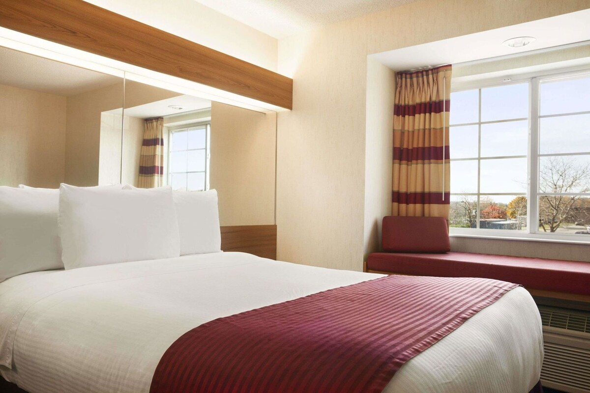 A hotel room with a large bed, a window with curtains, a bench, and a mirror.