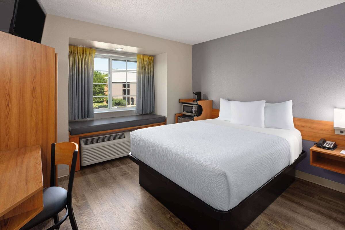 A hotel room with a bed, a desk, a chair, a heater, a window with curtains, and a small table with a phone.