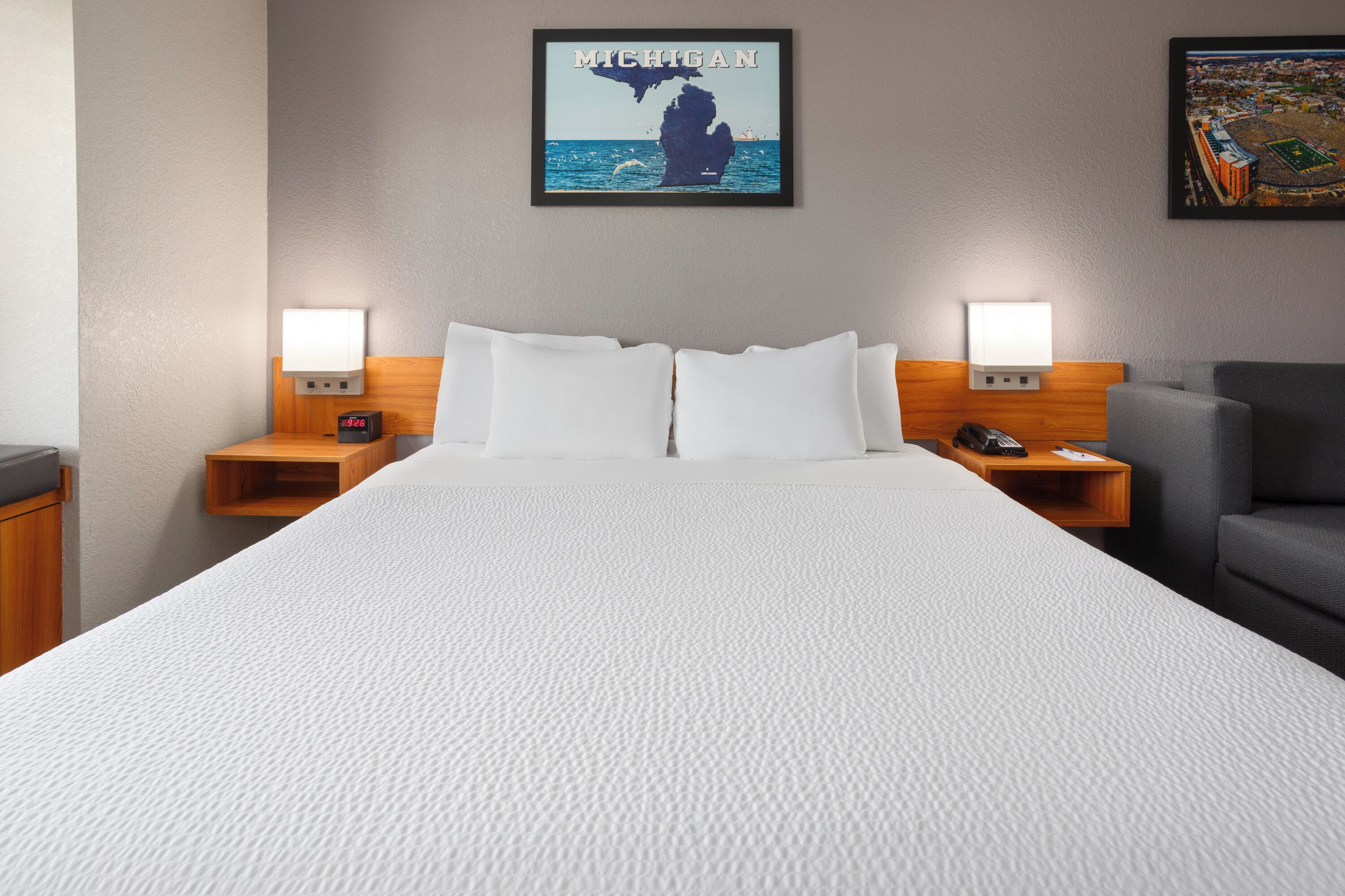 A hotel room with a large bed, two bedside tables with lamps, a gray armchair, and a framed picture of Michigan on the wall.
