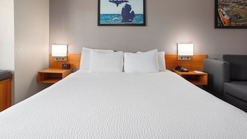 A hotel room with a large bed, two bedside tables with lamps, a gray armchair, and a framed picture of Michigan on the wall.