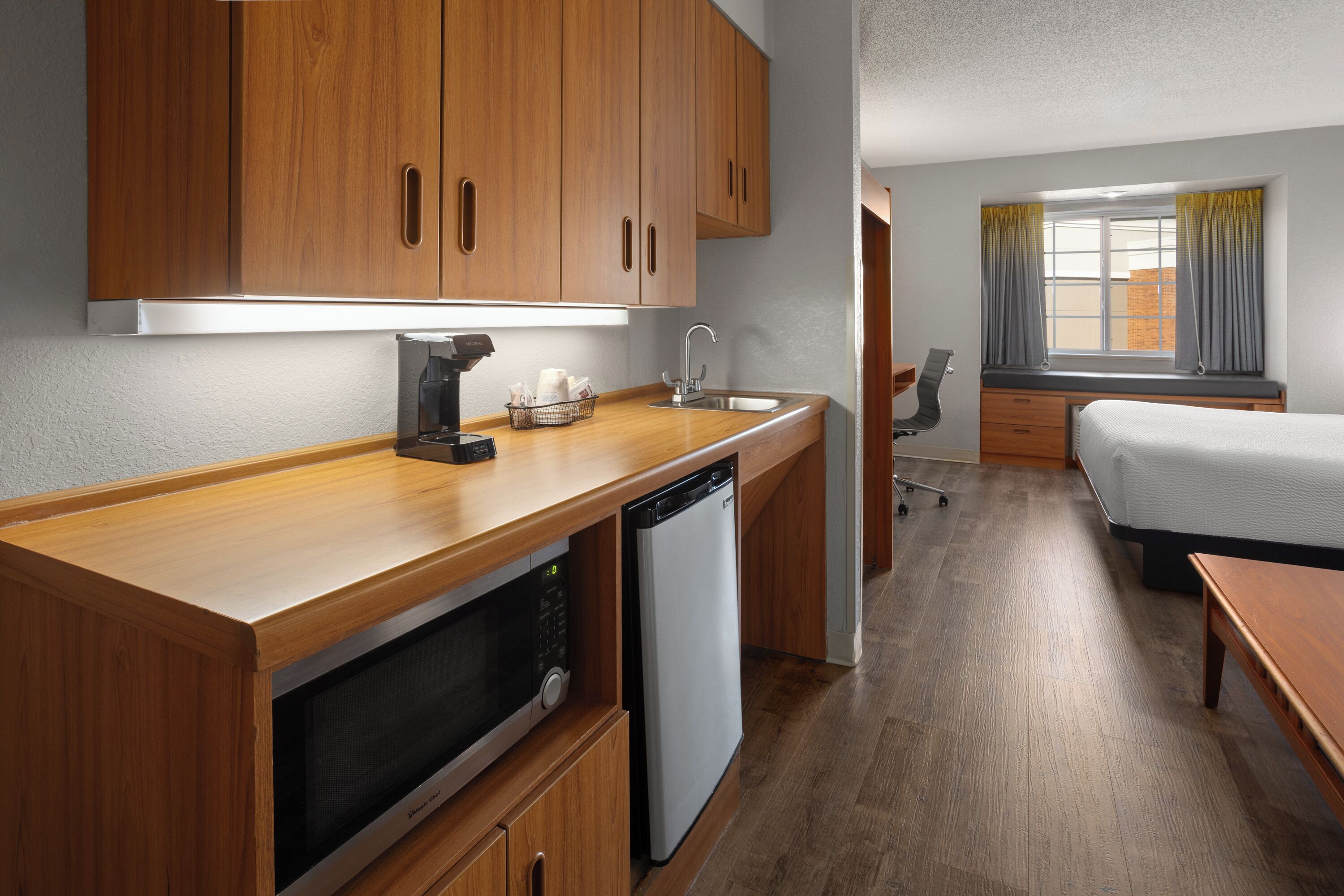 A compact hotel room with wooden cabinetry, a kitchenette, a bed, and a window with curtains.