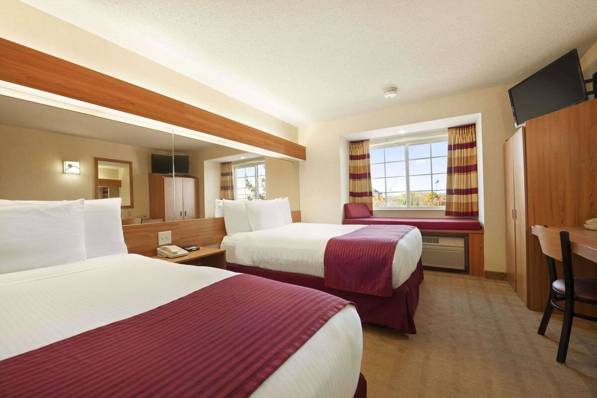 A hotel room with two beds, a desk, a chair, a TV, a window with curtains, and a mirror.