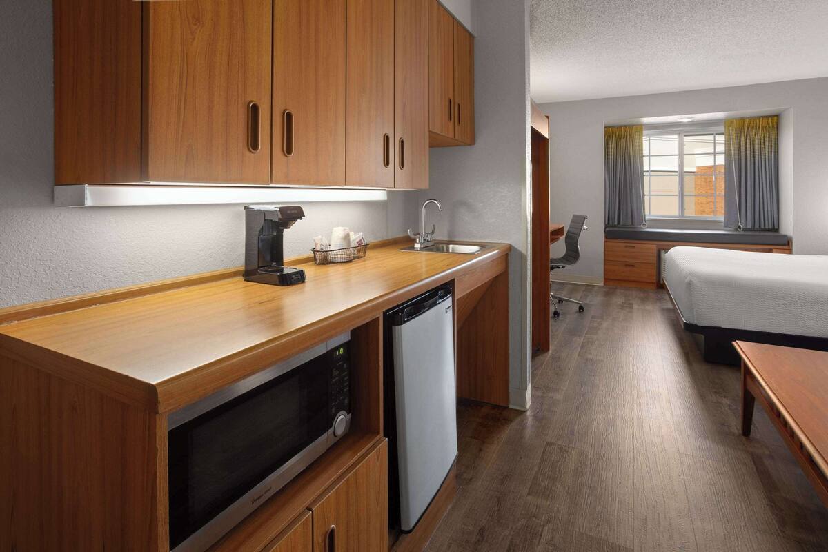 A compact hotel room with a kitchenette, a bed, a desk, and a window with curtains.