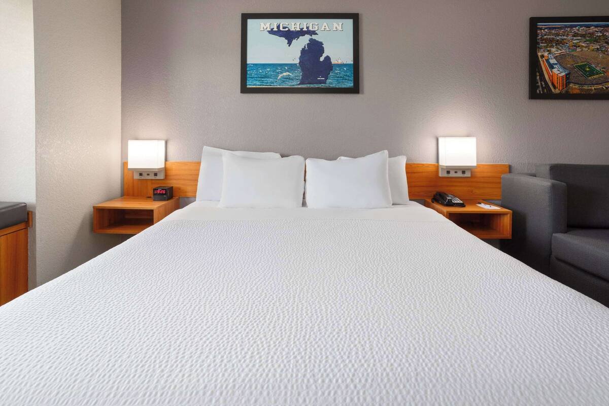 A hotel room with a large bed, two bedside tables with lamps, a gray armchair, and a wall-mounted picture of Michigan.