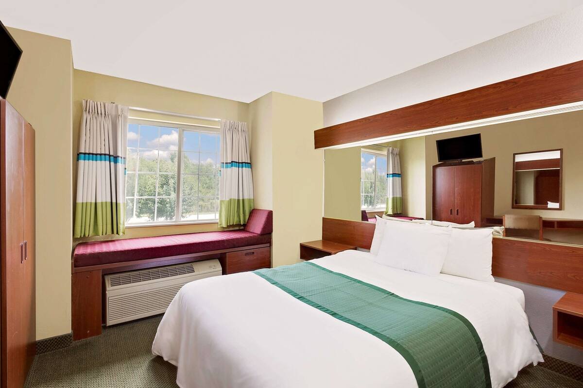 A hotel room with a bed, a window with curtains, a bench, a desk, a mirror, and a television.