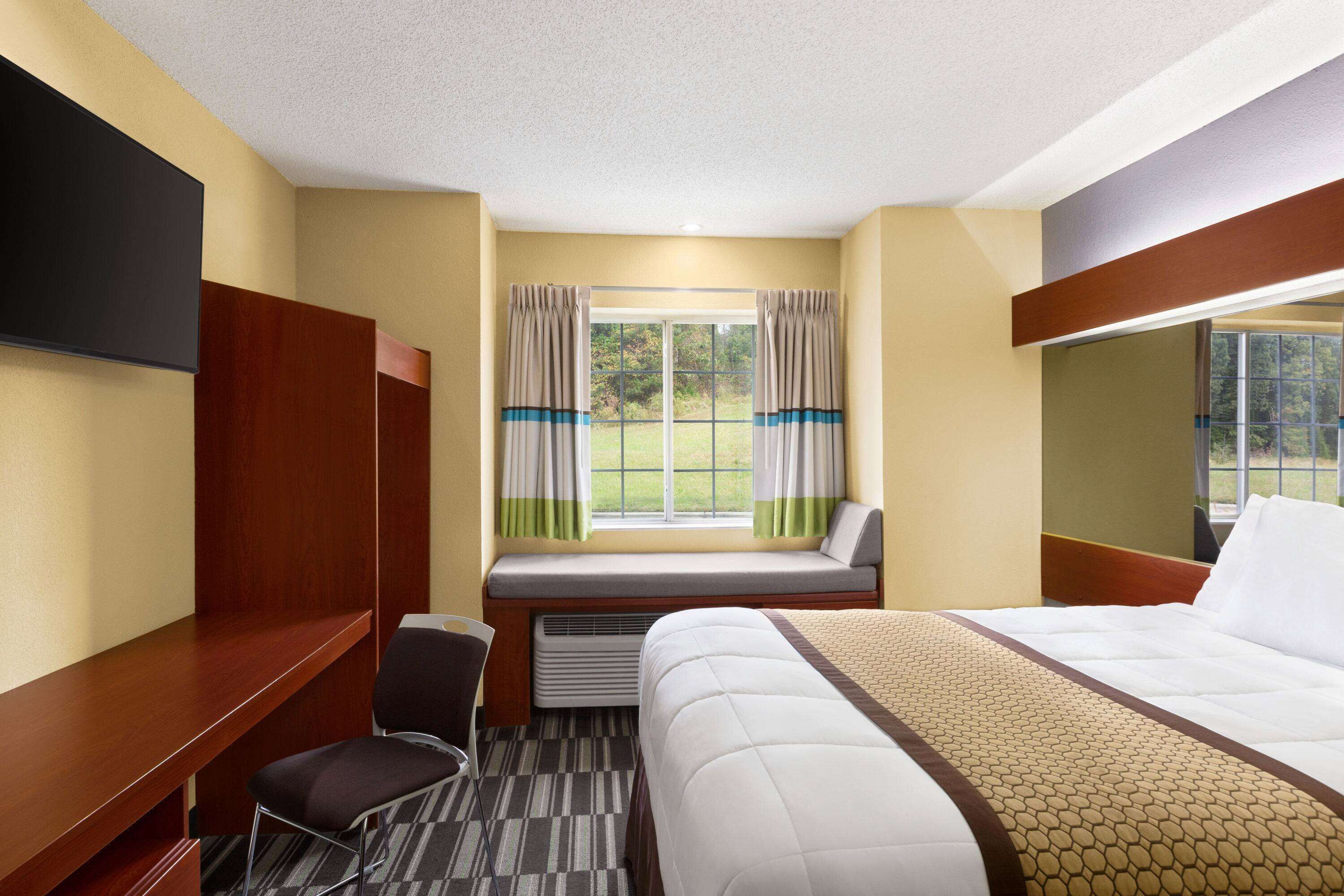 Une chambre d’hôtel avec un lit, un bureau, une chaise, une fenêtre avec des rideaux et un téléviseur fixé au mur.