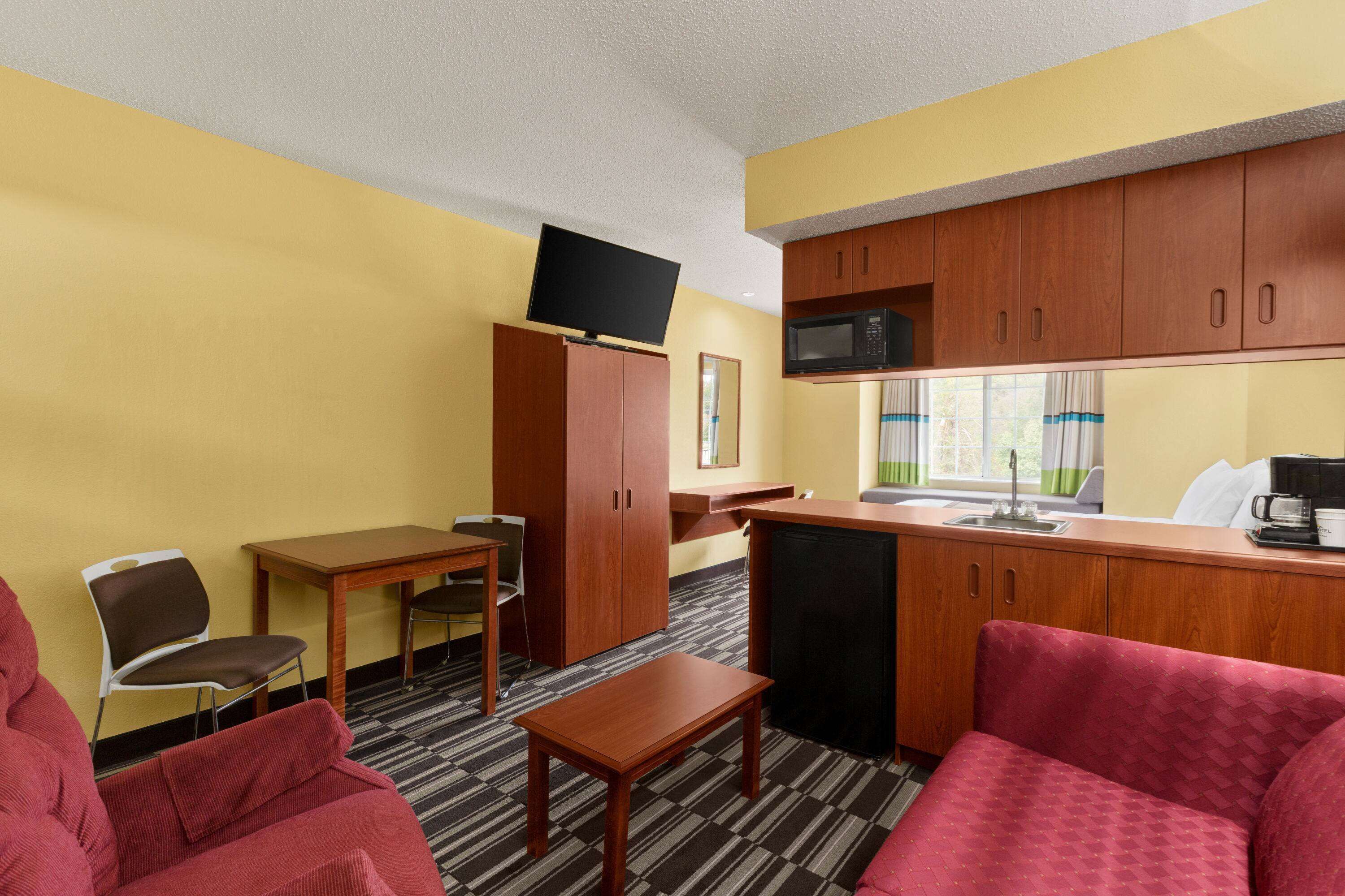 A compact hotel room with a kitchenette, a sofa, a small table, and a TV mounted on the wall.