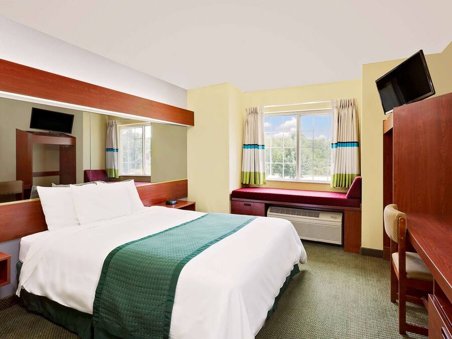 Una habitación de hotel con cama, escritorio, silla, televisor y ventana con cortinas.
