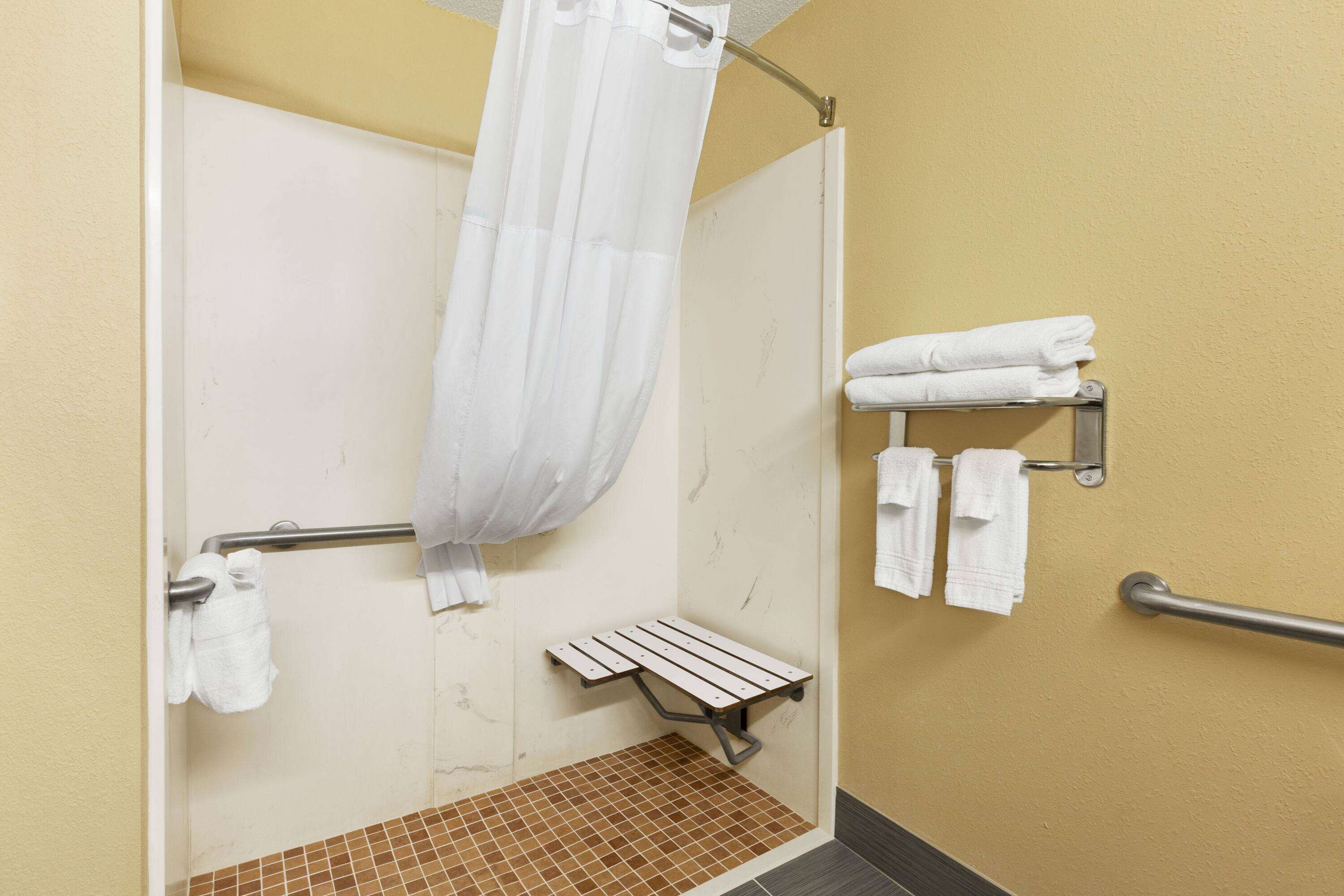 A shower area with a white shower curtain, a grab bar, a bench, and towels on a rack.