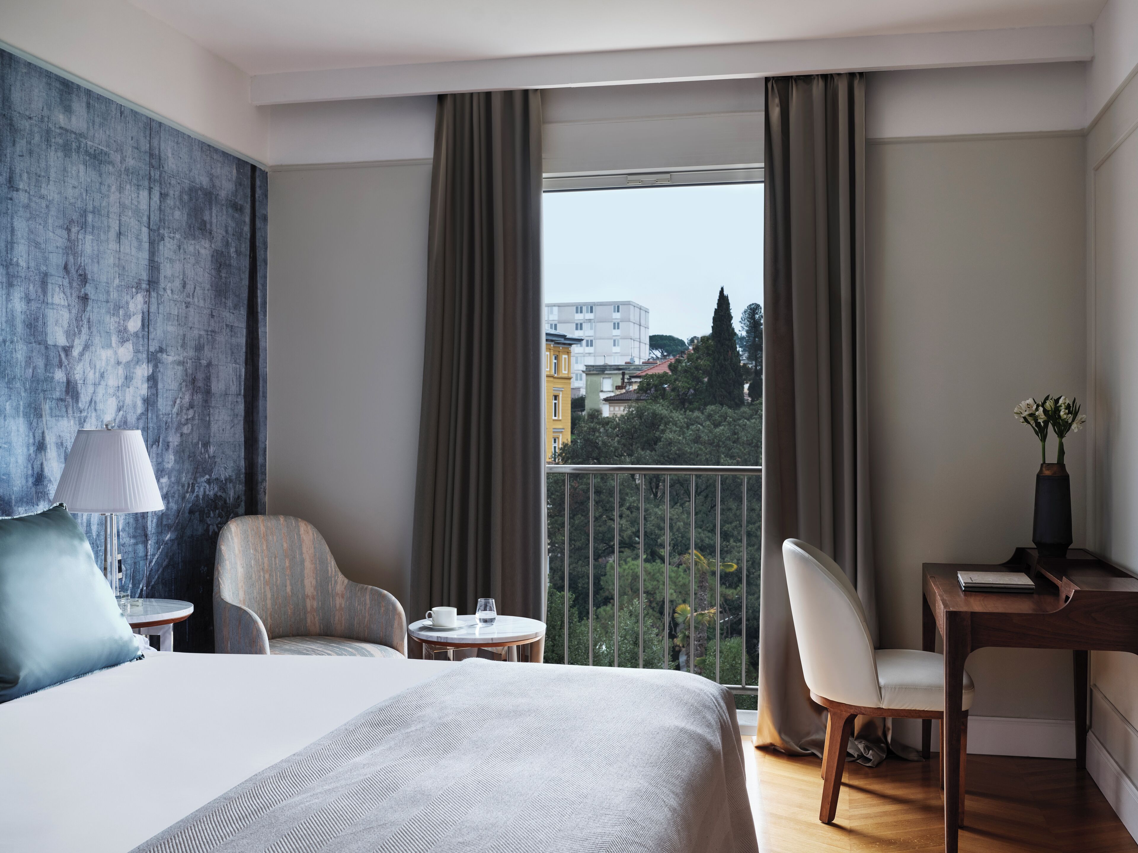 A hotel room with a large bed, a desk, two chairs, a small table with a cup and glass, and a view of buildings and trees through the window.