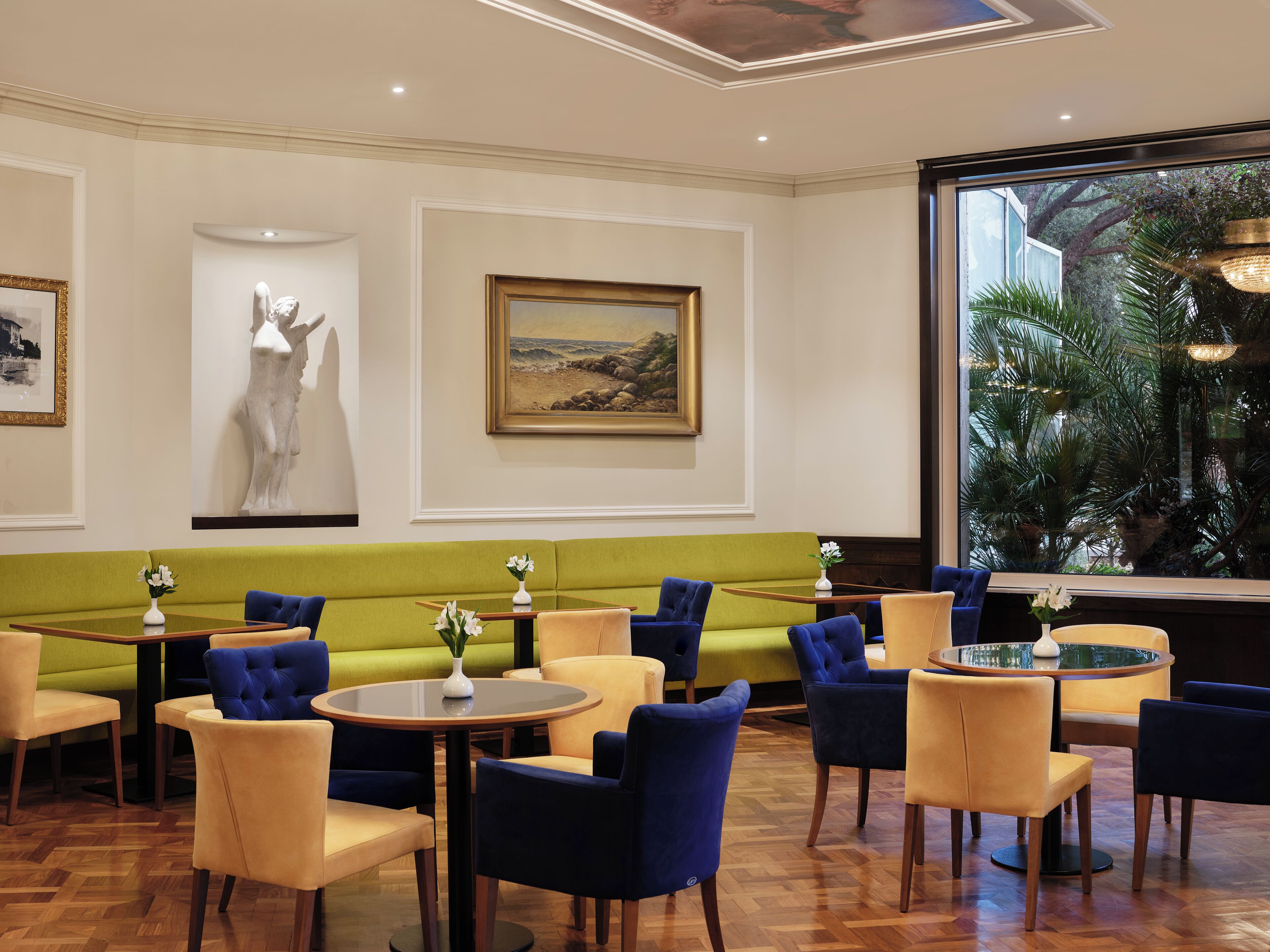 Un espace intérieur bien éclairé, avec un canapé vert, des chaises bleues et jaunes, et une table ronde agrémentée d'un vase de fleurs.