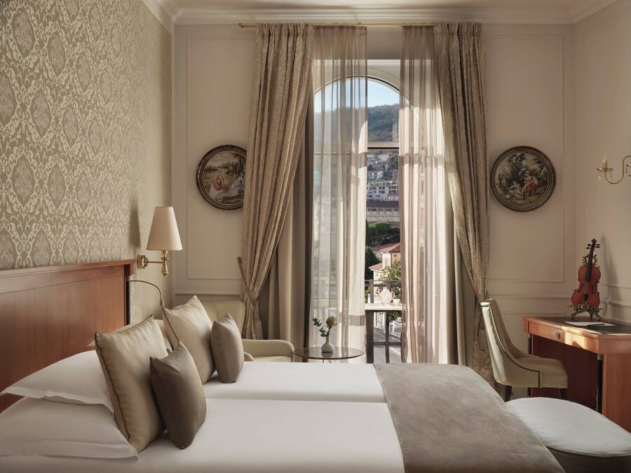 Ein Hotelzimmer mit einem großen Bett, einem Schreibtisch, einem Sessel und Blick auf die Stadt durch transparente Vorhänge.