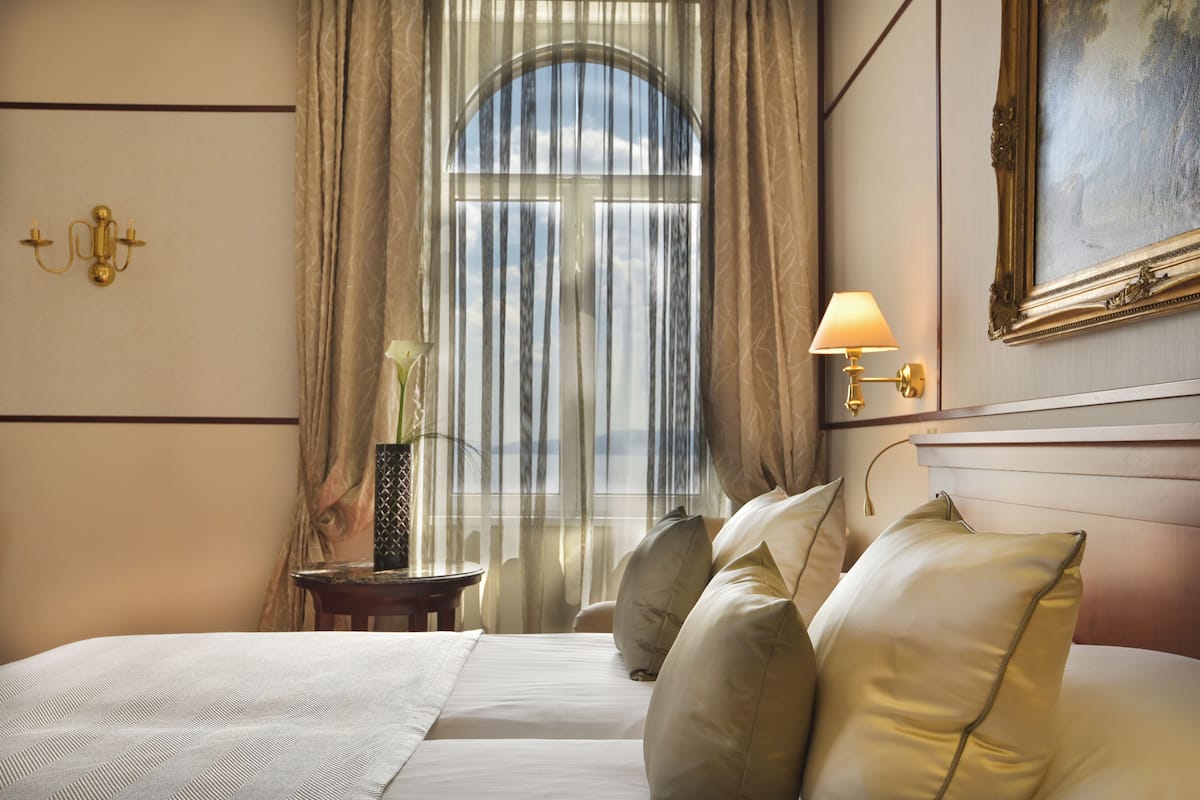 Habitación de hotel con una cama grande, una mesita de noche con lámpara, un cuadro en la pared y vistas a través de cortinas traslúcidas.