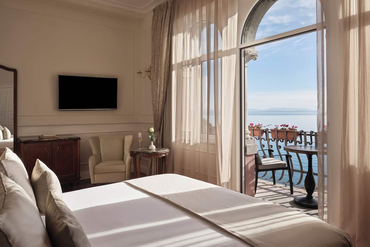 Habitación de hotel con una cama grande, un televisor, una mesita con un florero y vistas al mar a través de un ventanal amplio.