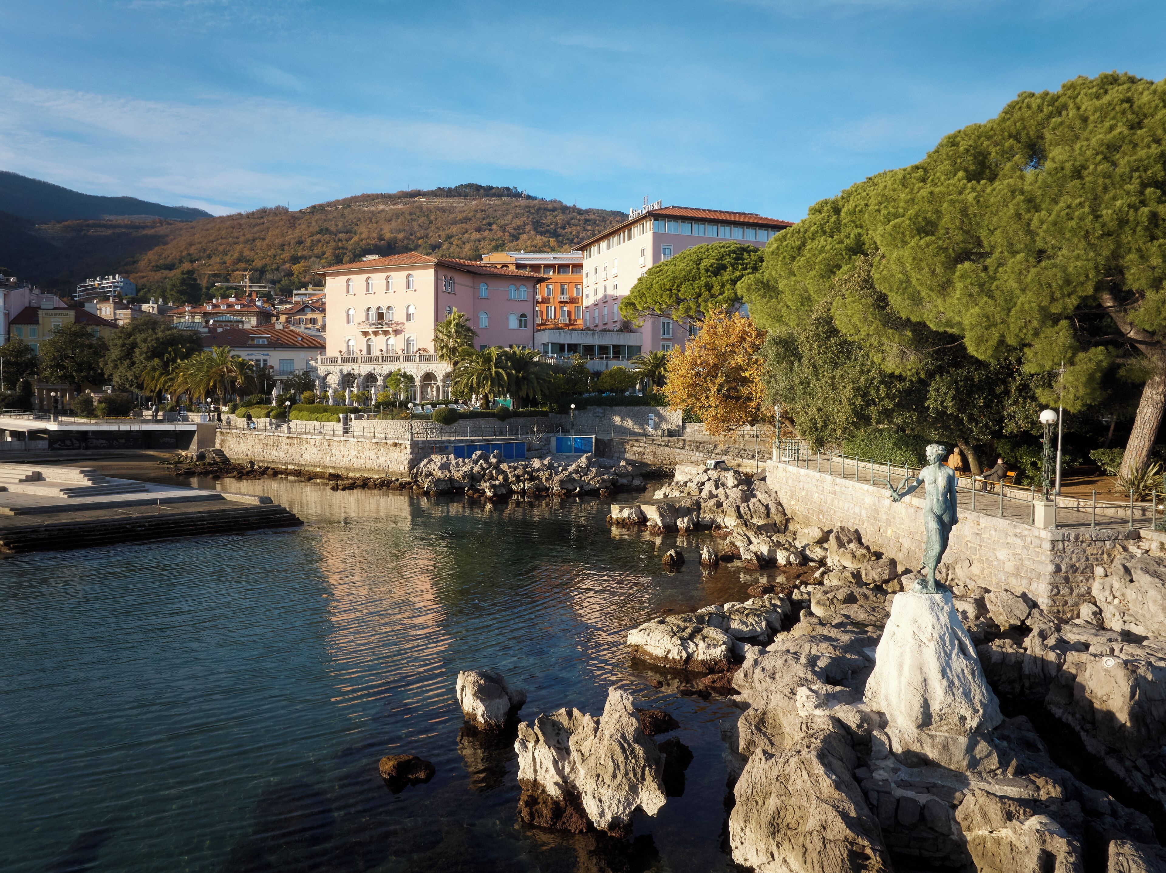 Una zona ribereña con una estatua, costa rocosa y edificios al fondo.