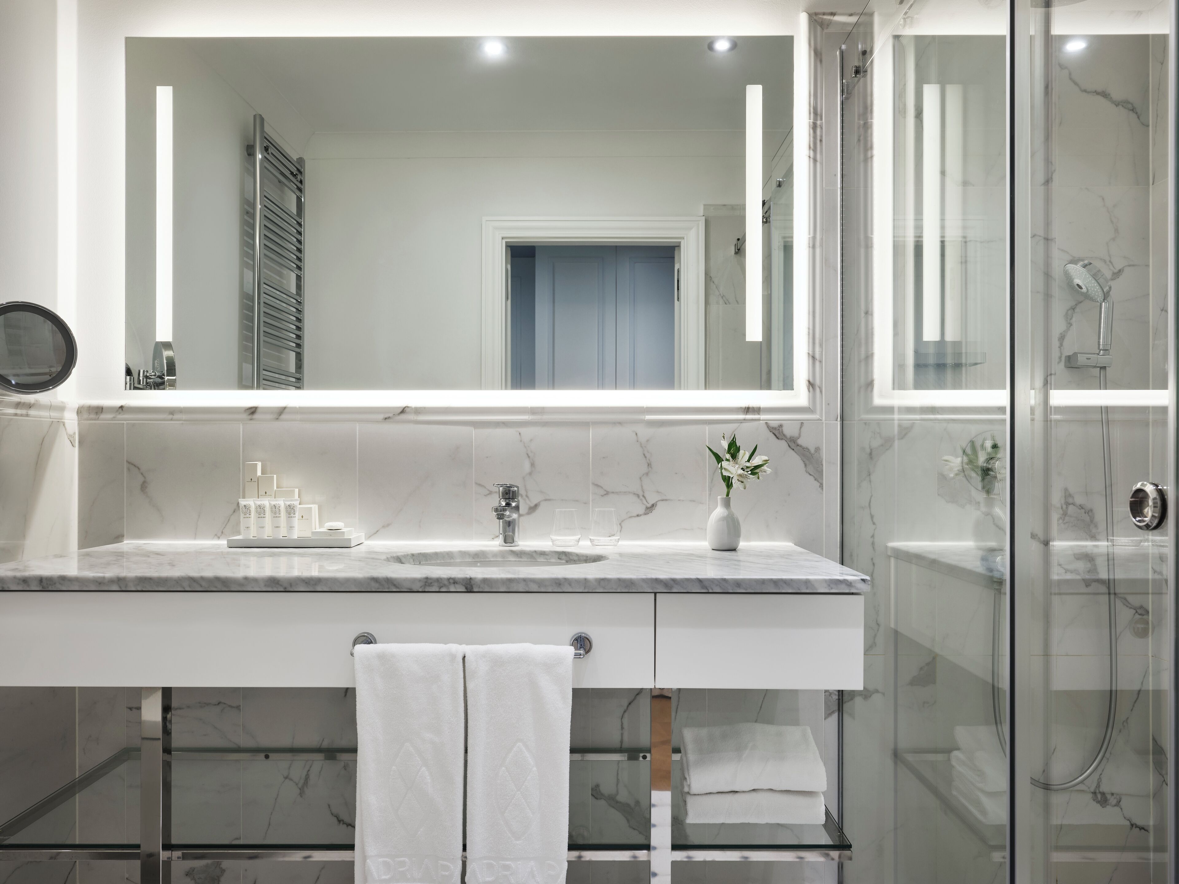 A modern bathroom with a large mirror, marble countertops, a glass shower enclosure, and a towel rack.