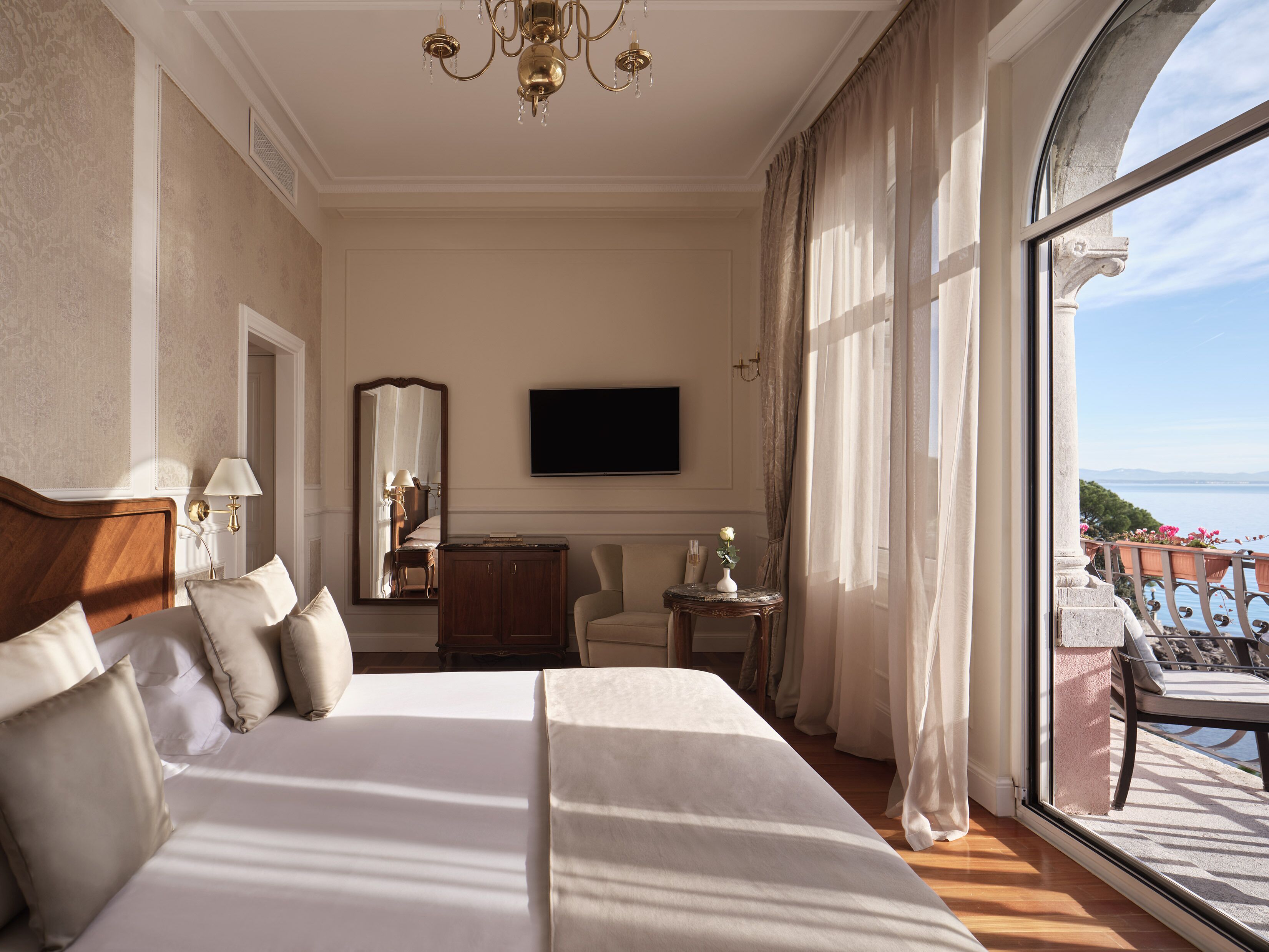 Quarto de hotel com uma cama grande, televisão, espelho, mesa pequena com um vaso e varanda com vista.