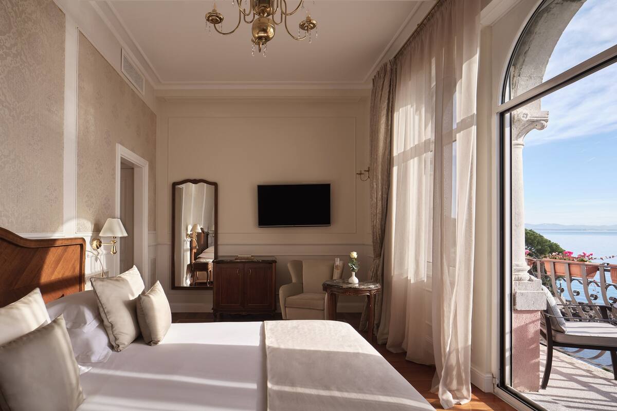 Habitación de hotel con una cama grande, televisión, espejo, una mesita con un florero y un balcón con vistas.