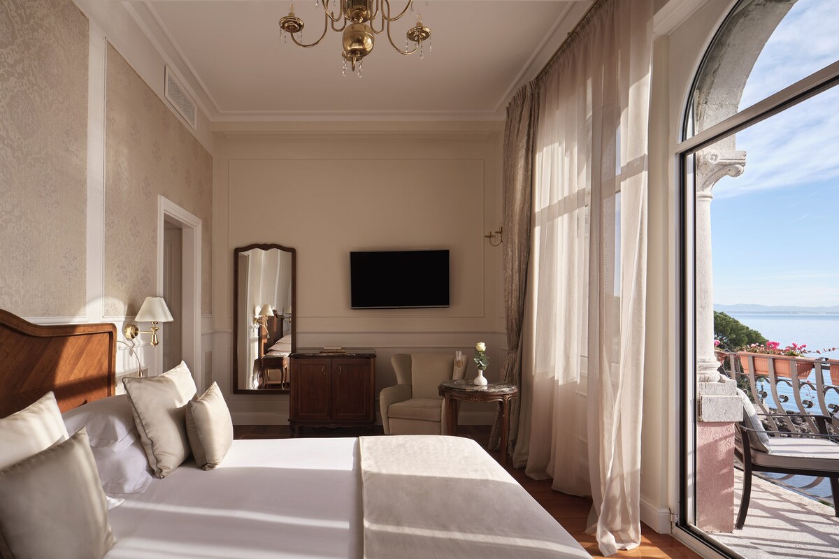 Une chambre d’hôtel avec un grand lit, une télévision, un miroir, une petite table avec un vase et un balcon avec vue.