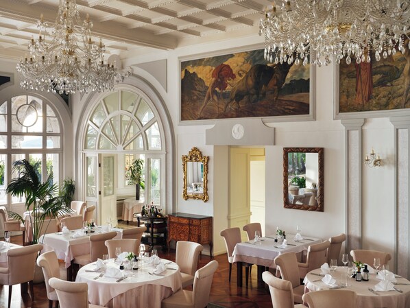 Un comedor formal con varias mesas dispuestas para el servicio, ventanas amplias y candelabros ornamentados.