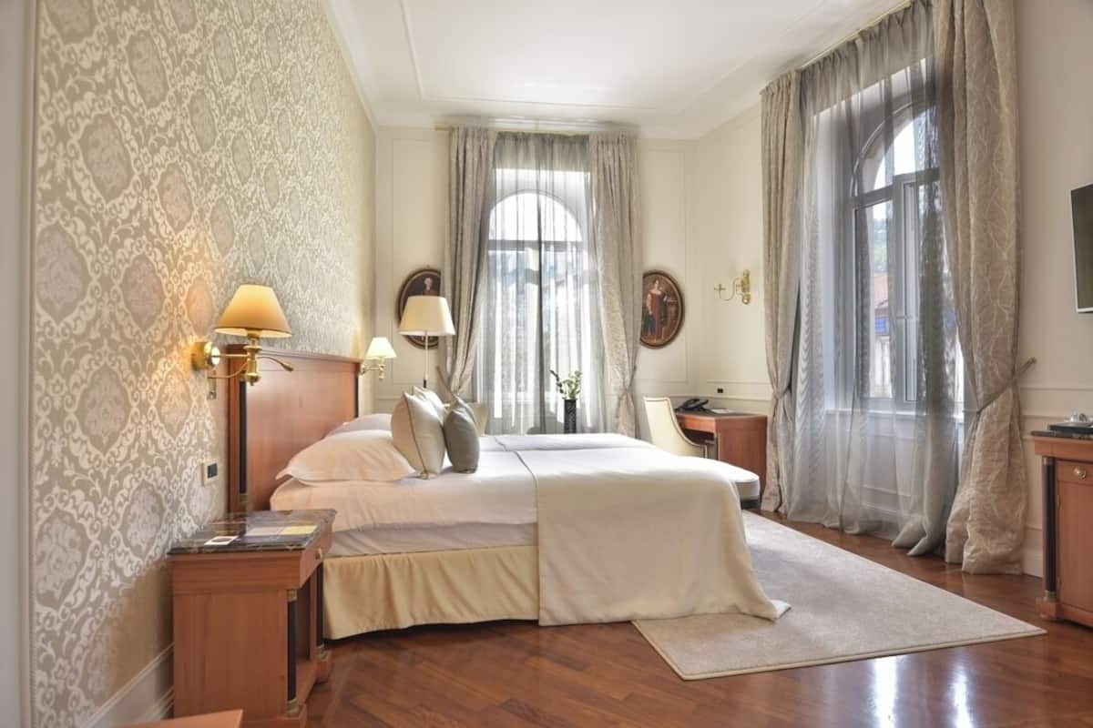 Un dormitorio con cama, mesitas de noche, un escritorio, una silla y un ventanal con cortinas.