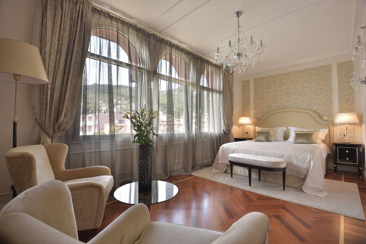 Habitación de hotel con una cama grande, dos sillones, una mesa de centro redonda, un banco y vistas a la ciudad a través de amplias ventanas.
