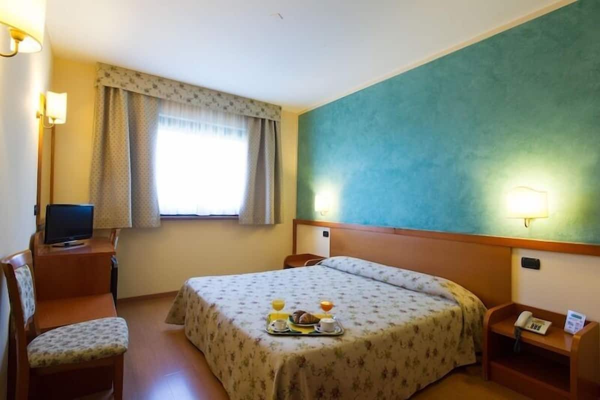 A hotel room with a bed, a desk with a computer, a chair, a bedside table with a phone, and a tray with breakfast items.