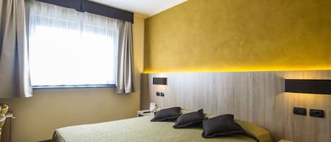 A hotel room with a bed, a bedside table, a lamp, a window with curtains, and a wall-mounted light fixture.