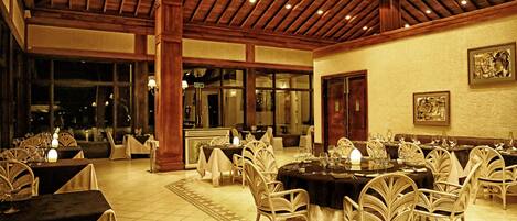 A restaurant with round tables and wicker chairs, wooden ceiling beams, and large windows.