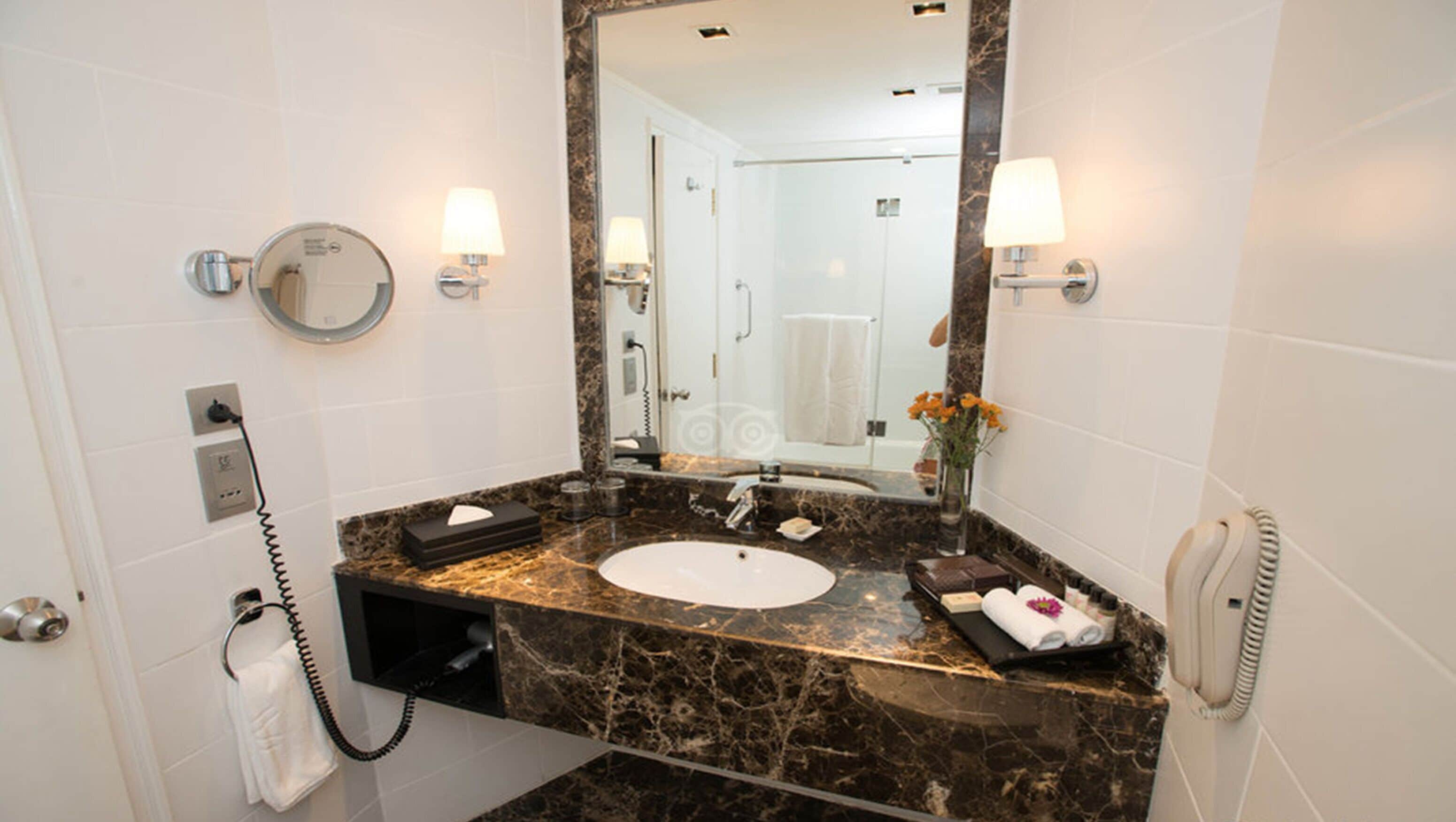 Un baño con encimera de mármol, lavamanos, espejo, toallero, teléfono y un florero con flores.