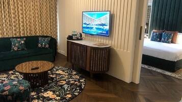 Una habitación de hotel con un sofá verde, una mesa de centro redonda, un televisor montado en la pared y una cama con ropa de cama estampada con motivos florales.