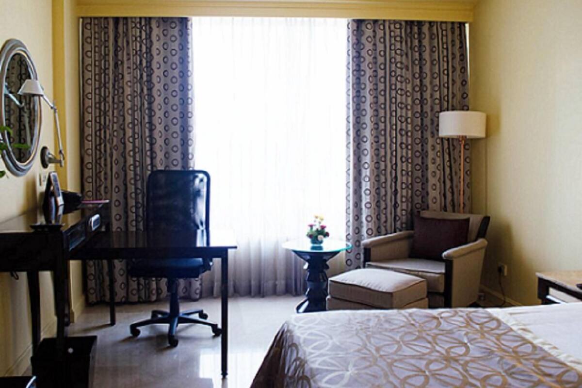 A hotel room with a bed, a desk, a chair, a small table with a lamp, and curtains.