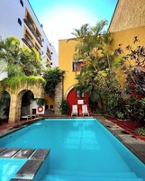 Una piscina con sillas de descanso, rodeada de plantas tropicales y un edificio de fachada amarilla.