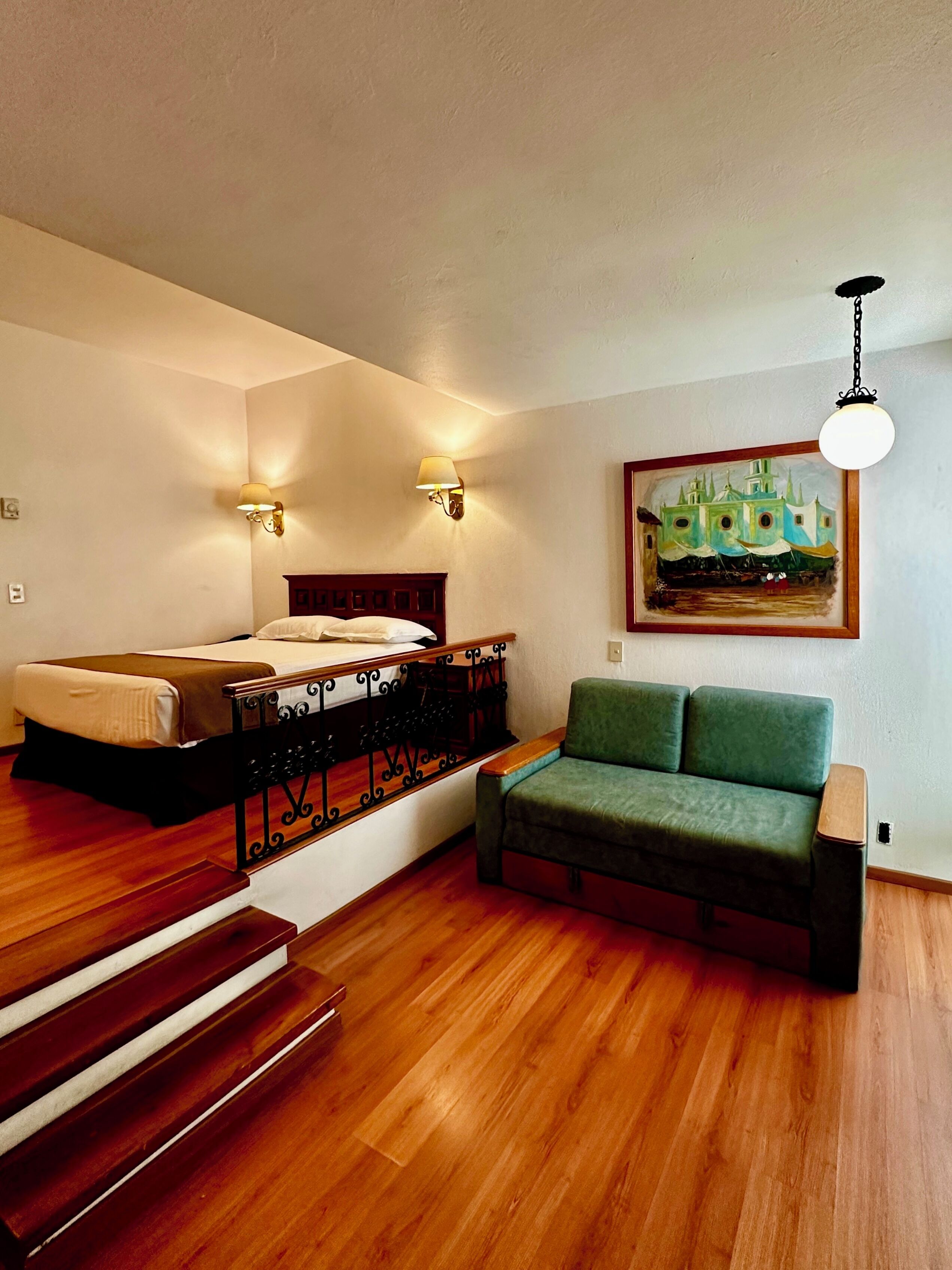 A bedroom with a bed, a green sofa, wooden flooring, a painting, and two wall-mounted lamps.