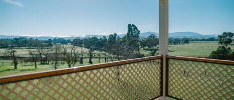 A covered balcony with a lattice railing overlooking a grassy field and distant hills.