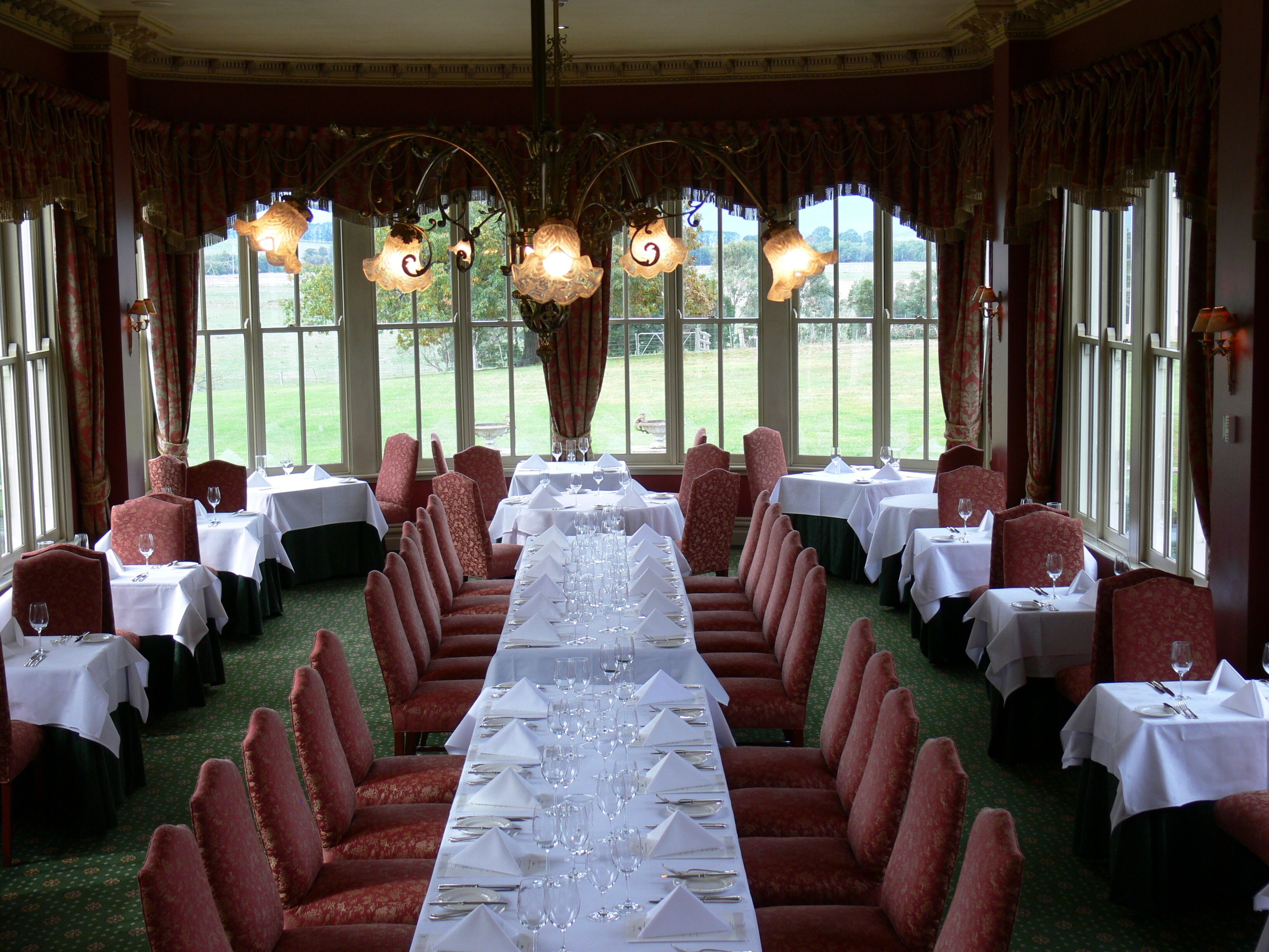 Een formele eetzaal met lange tafels gedekt voor een maaltijd, rode gestoffeerde stoelen en grote ramen met gordijnen.