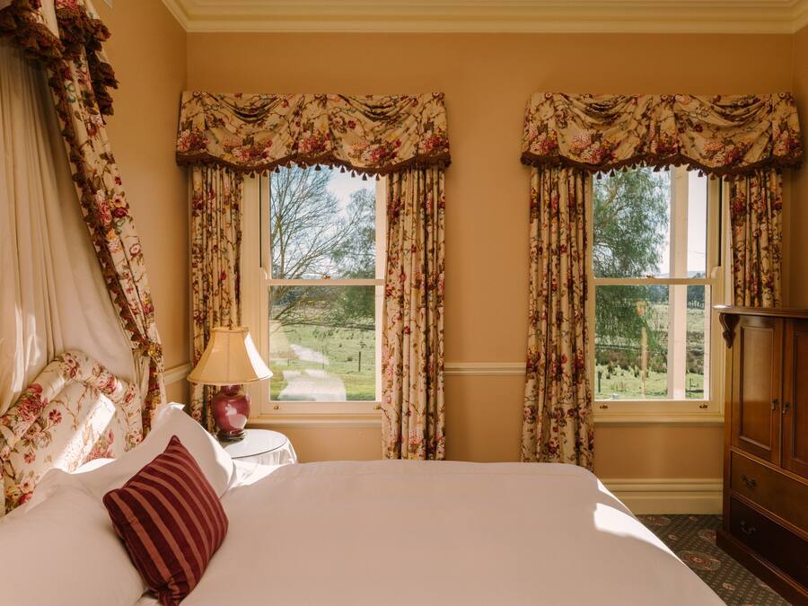 A bedroom with a large bed, floral curtains, a bedside lamp, and a view of the outdoors.