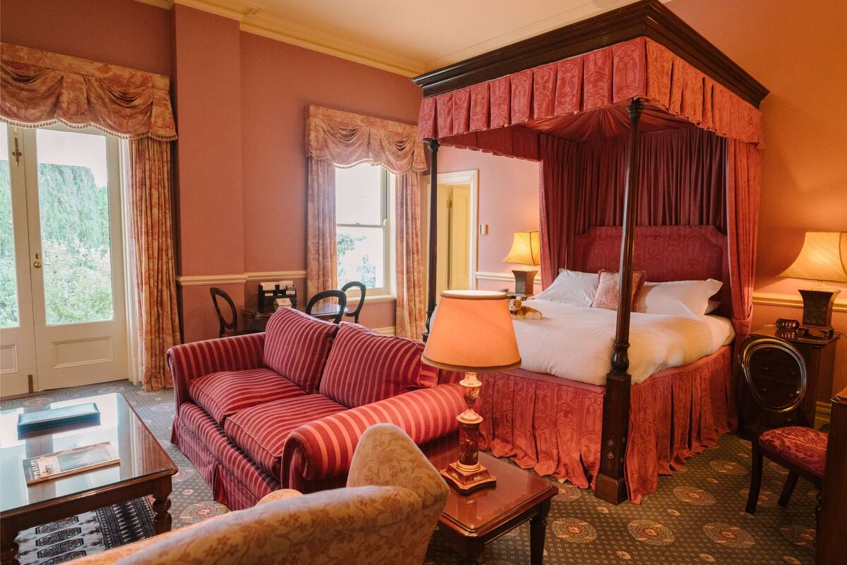 A four-poster canopy bed with a red and white bedspread, a red-striped sofa, a small table with a lamp, and a chair.