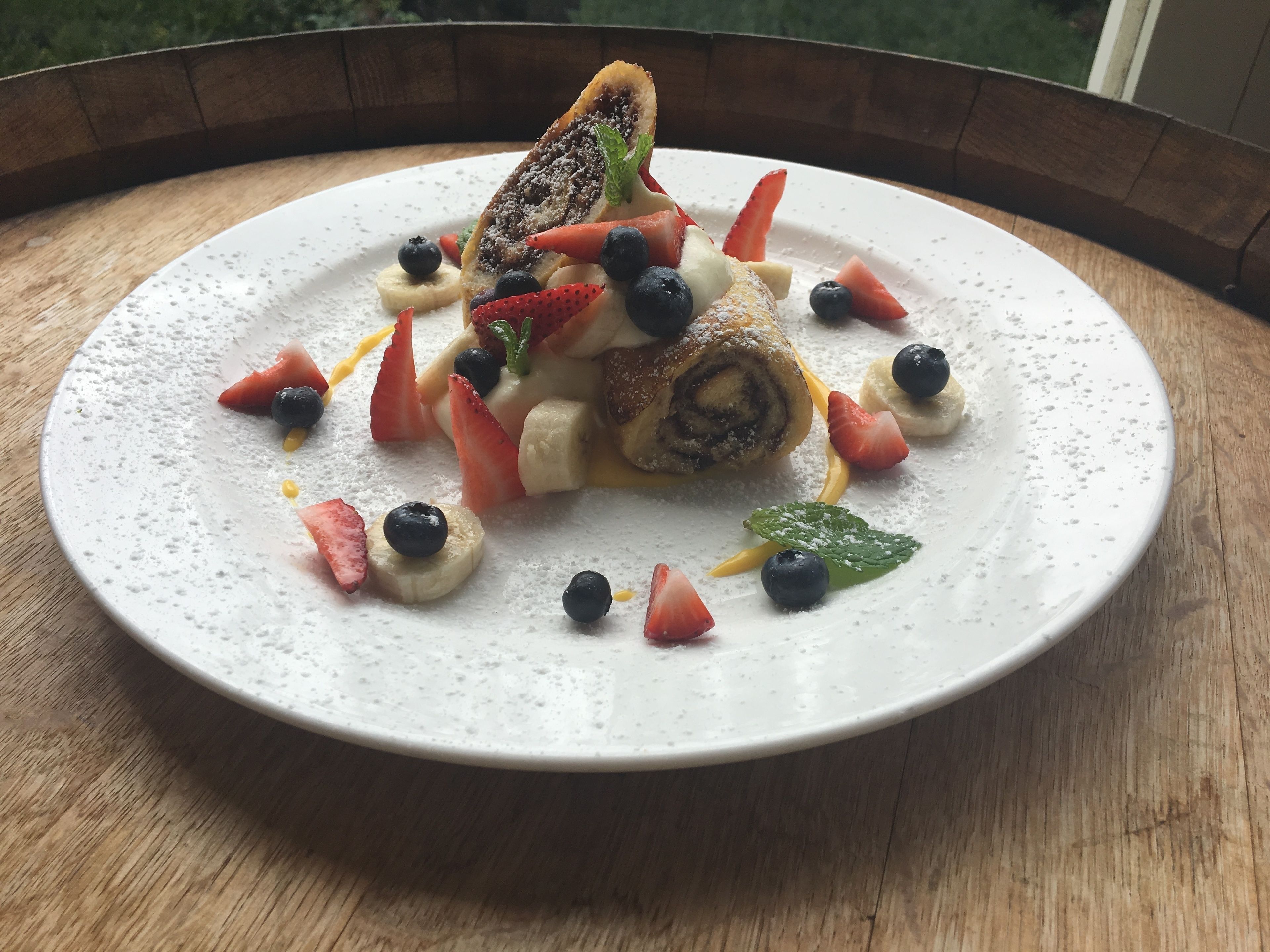 A dessert plate with a rolled pastry, strawberries, blueberries, banana slices, and mint leaves.