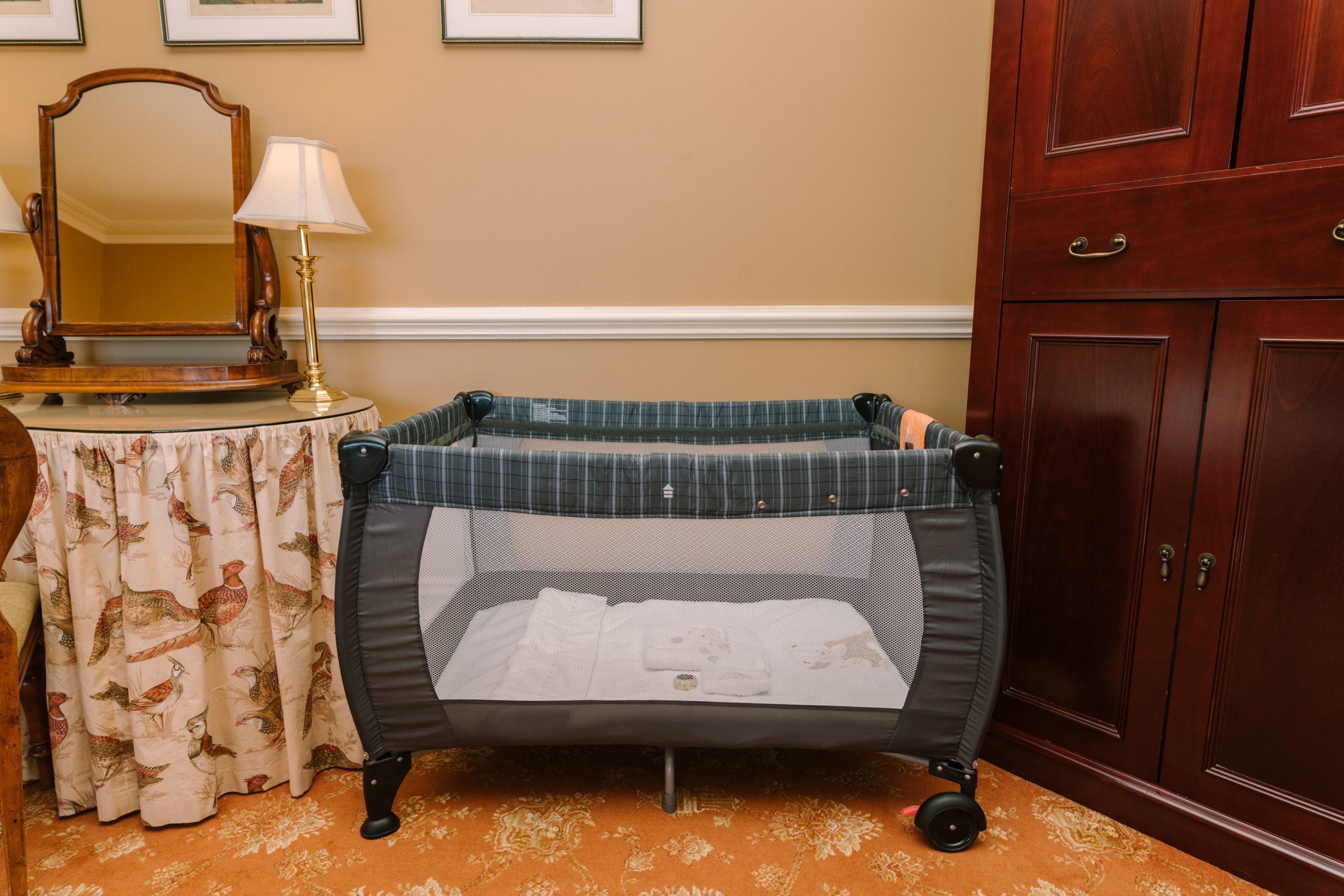 Una cuna para bebé con cubierta de malla, ubicada junto a una mesa con mantel floral y un espejo.
