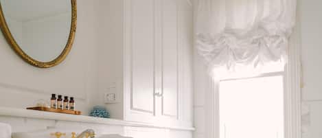 A bathroom with a large white sink, a pedestal tub, and a toilet. There is a window with a white curtain, and a mirror with a gold frame.